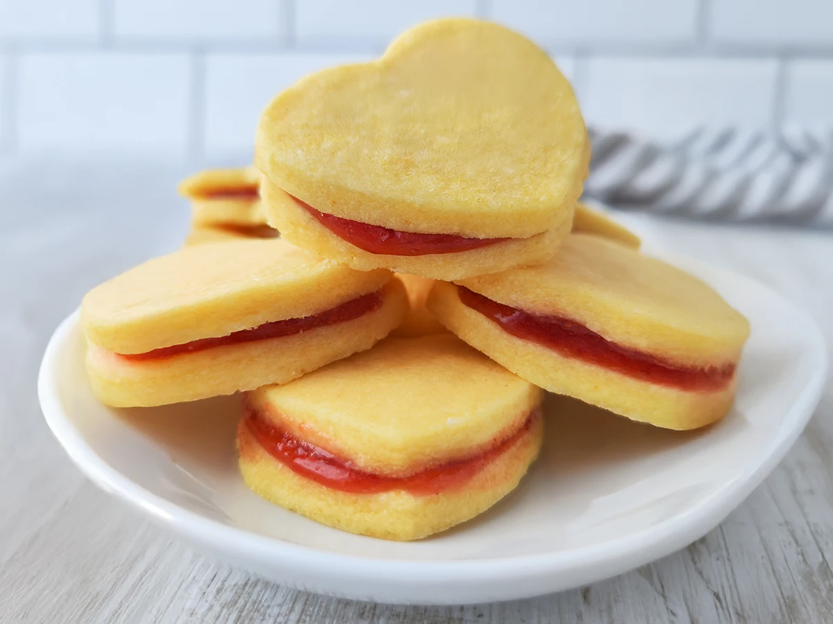 Ein Teller mit mehreren herzförmigen veganen Vanilleplätzchen, die mit roter Marmelade gefüllt sind.
