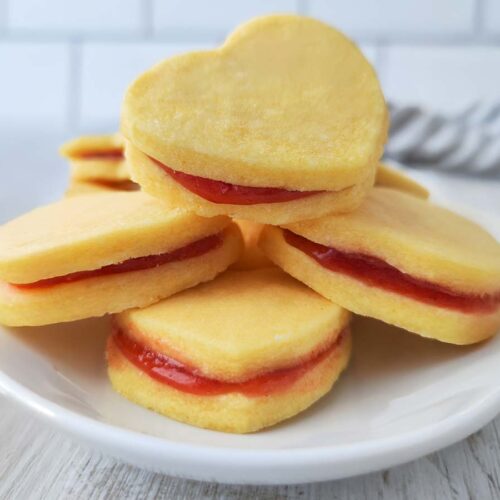 Mehrere gestapelte herzförmige vegane Vanilleplätzchen mit roter Marmelade auf einem weißen Teller.