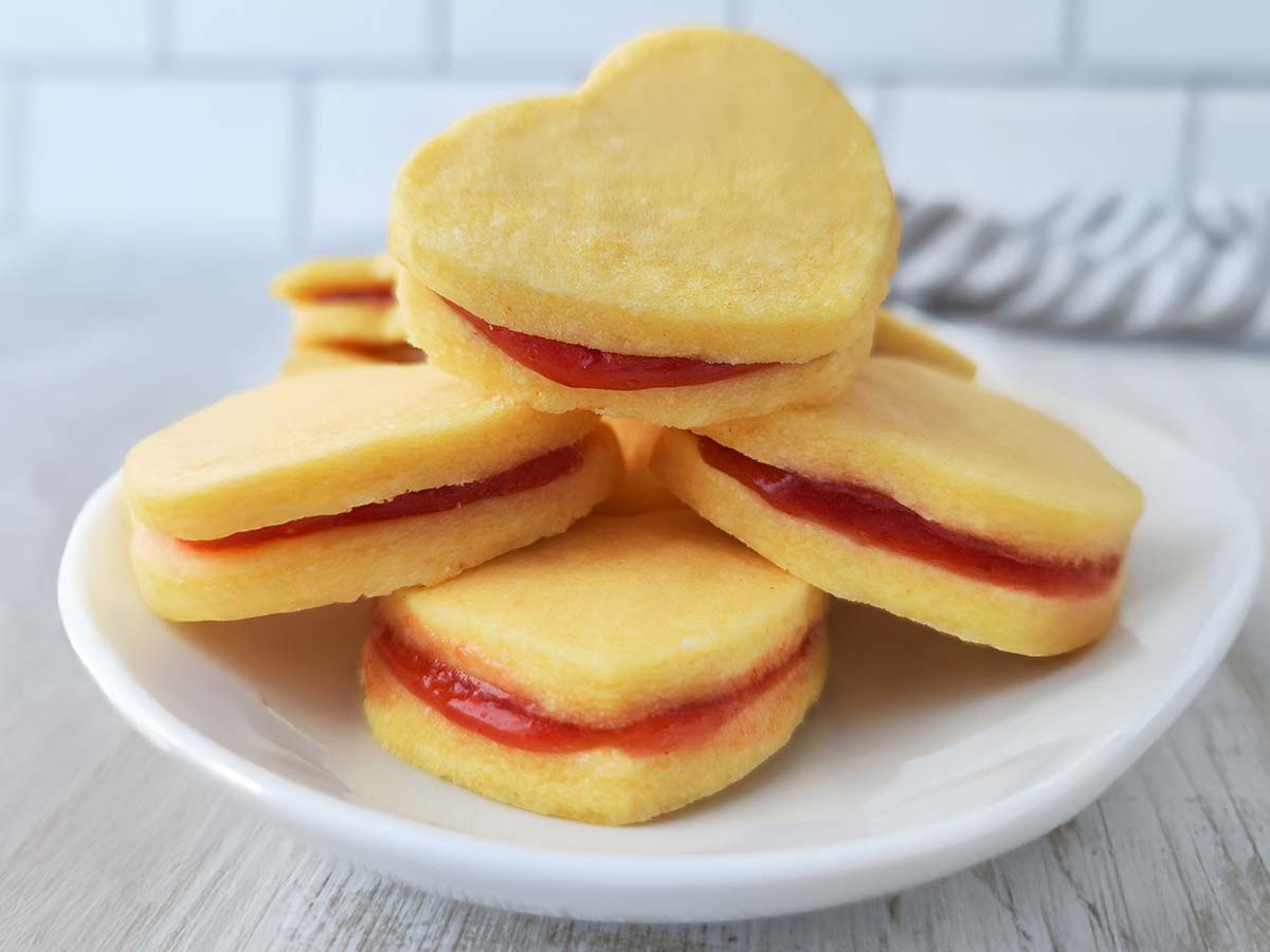 Mehrere gestapelte herzförmige vegane Vanilleplätzchen mit roter Marmelade auf einem weißen Teller.
