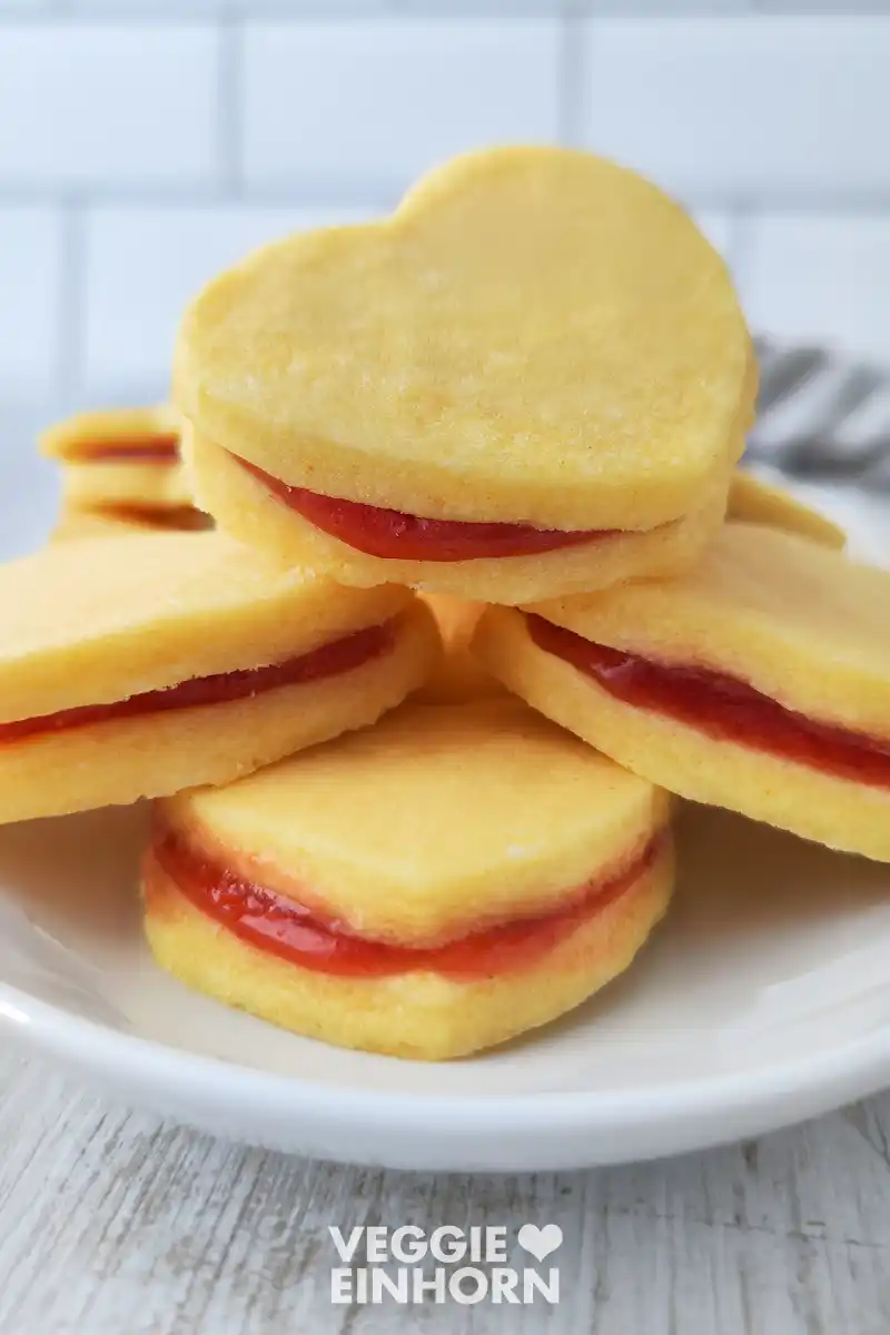 Gestapelte herzförmige vegane Vanilleplätzchen mit einer dünnen Schicht roter Marmelade dazwischen auf einem weißen Teller.