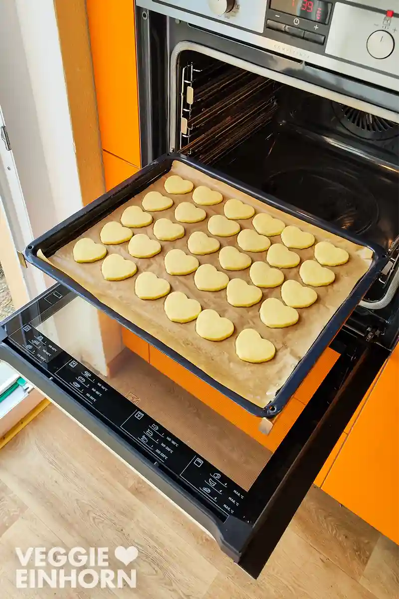 Ein Backblech mit ungefüllten herzförmigen veganen Vanilleplätzchen, aus dem geöffneten Backofen heraus gezogen.