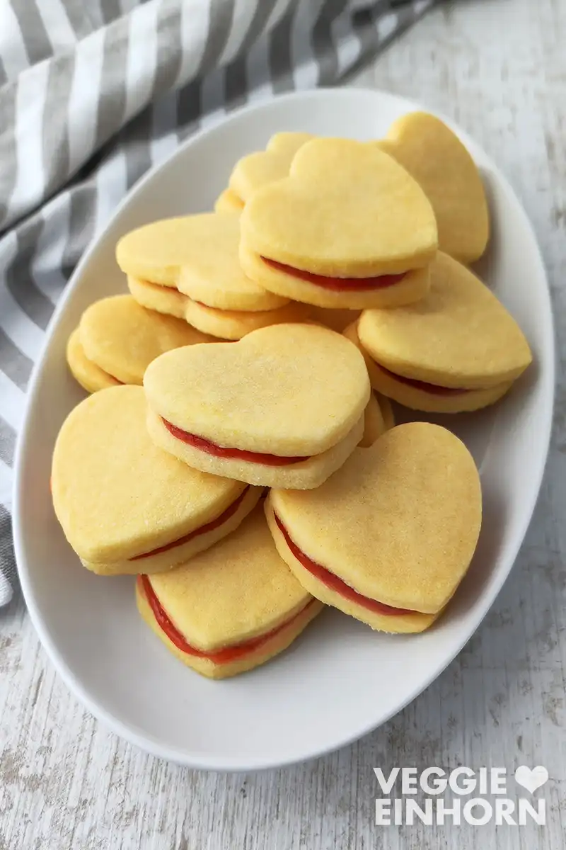 Ein weißer Plätzchenteller voller herzförmiger veganer Vanilleplätzchen mit roter Marmelade dazwischen.