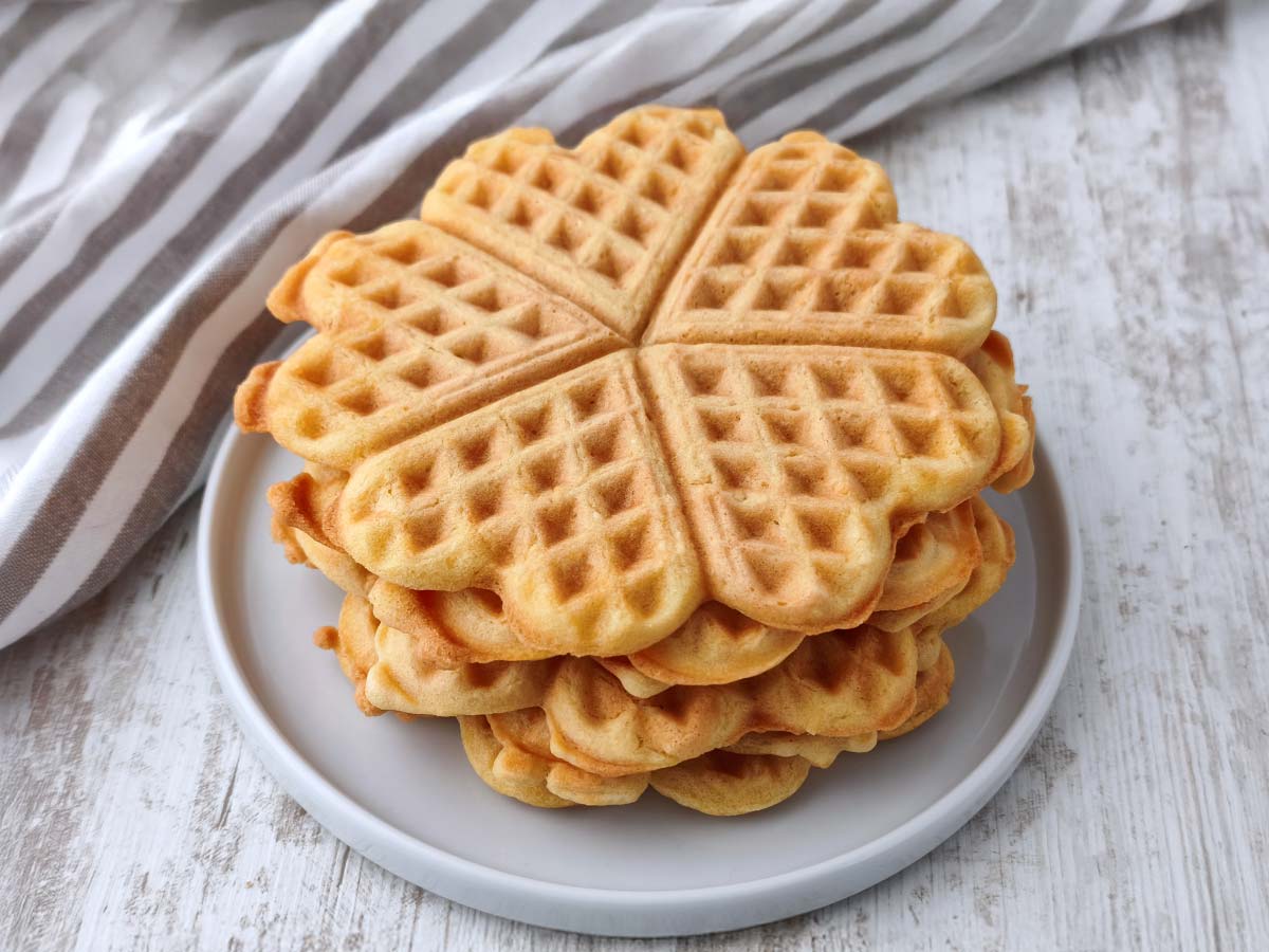 Ein Stapel goldbrauner, herzförmiger veganer Waffeln liegt auf einem grauen Teller vor einem hellen Holzuntergrund mit einem gestreiften Tuch im Hintergrund.