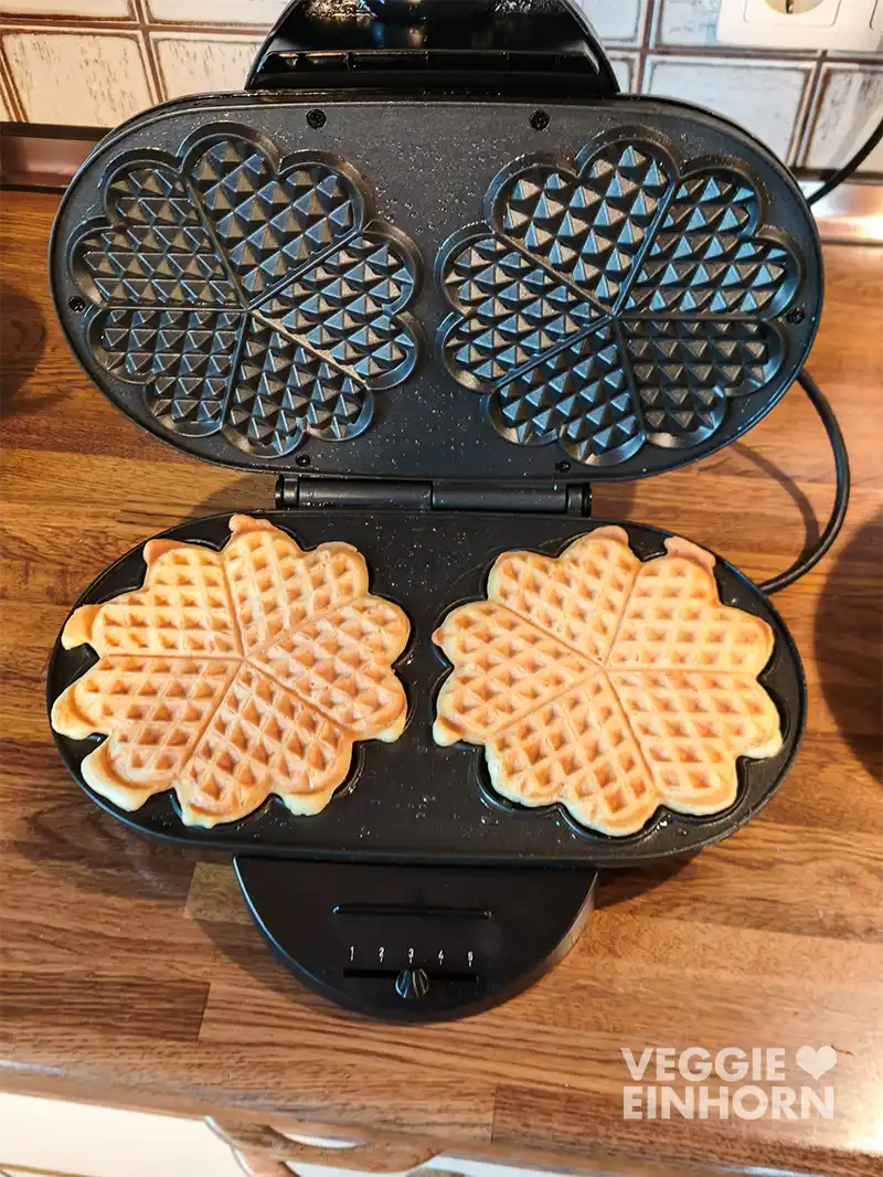 Ein geöffnetes schwarzes Waffeleisen mit zwei veganen Waffeln darin steht auf einer Holzarbeitsfläche.