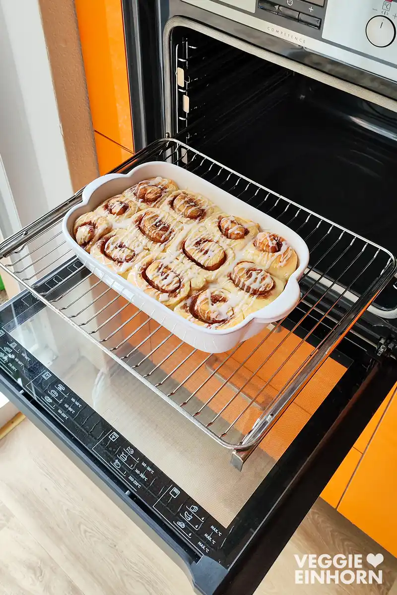 Vegane Zimtschnecken mit Zuckerguss in einer weißen Auflaufform im geöffneten Ofen.