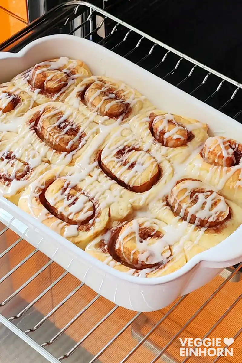 Nahaufnahme von gebackenen veganen Zimtschnecken mit Zuckerguss in einer weißen Form im Ofen.