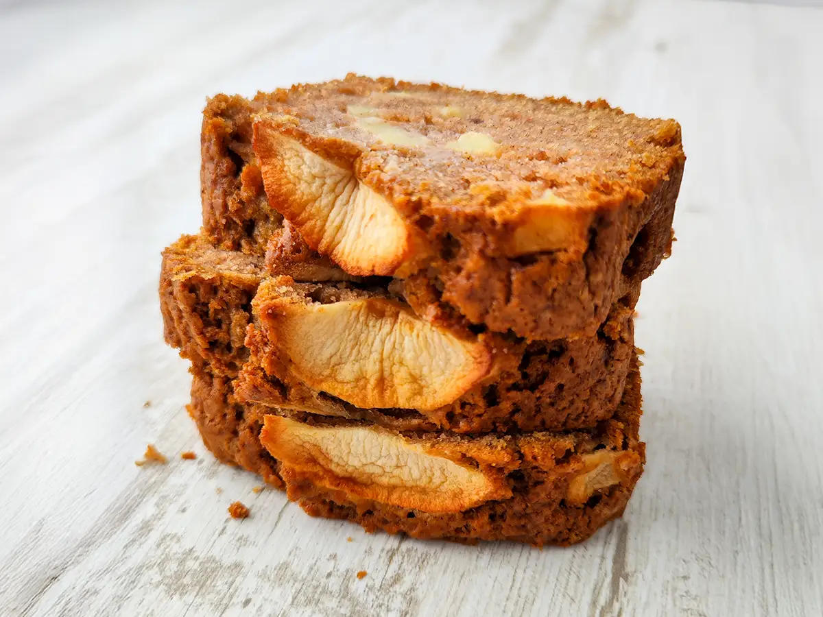 Drei Scheiben veganer Apfelkuchen, gestapelt aufeinander, mit sichtbaren Apfelstücken im Teig, auf einem hellen Holztisch.