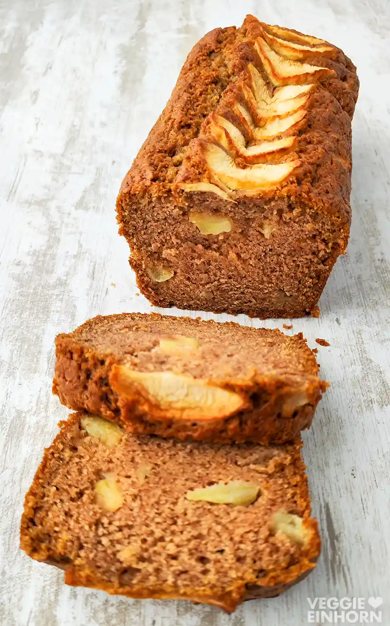 Veganer Apfelkuchen aus der Kastenform, angeschnitten und mit Apfelstücken im Teig, zwei Scheiben vor dem Kuchen liegend, auf hellem Holztisch fotografiert.