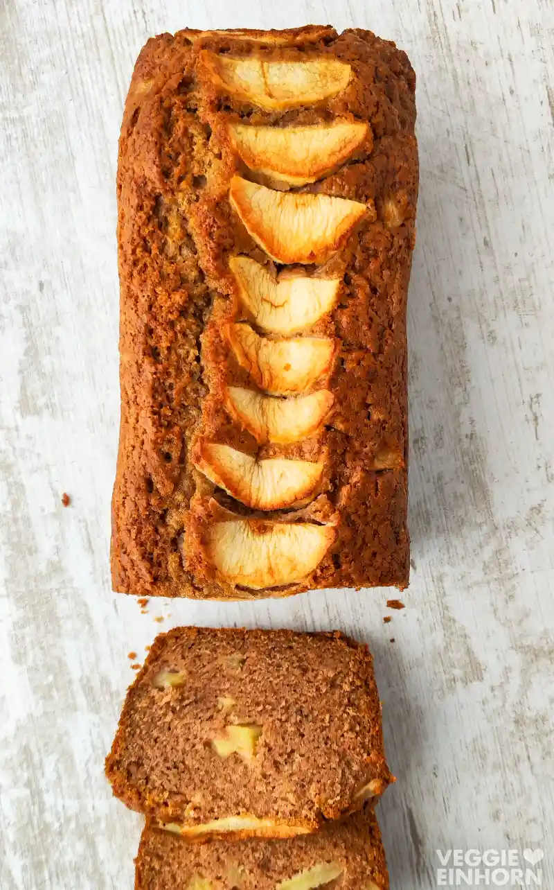 Veganer Apfelkuchen aus der Kastenform, von oben fotografiert, mit Apfelspalten auf der Oberfläche und angeschnittenen Scheiben auf hellem Holztisch.