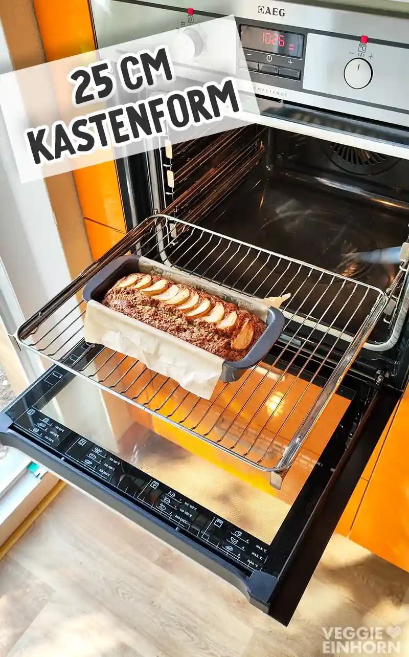 Ein veganer Apfelkuchen in einer 25 cm Kastenform, mit Backpapier ausgelegt und mit Apfelspalten belegt, steht im geöffneten Backofen auf dem Rost.