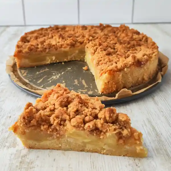 Ein Stück veganer Apfelkuchen mit Zimtstreuseln liegt vor einer Springform mit dem restlichen Kuchen.
