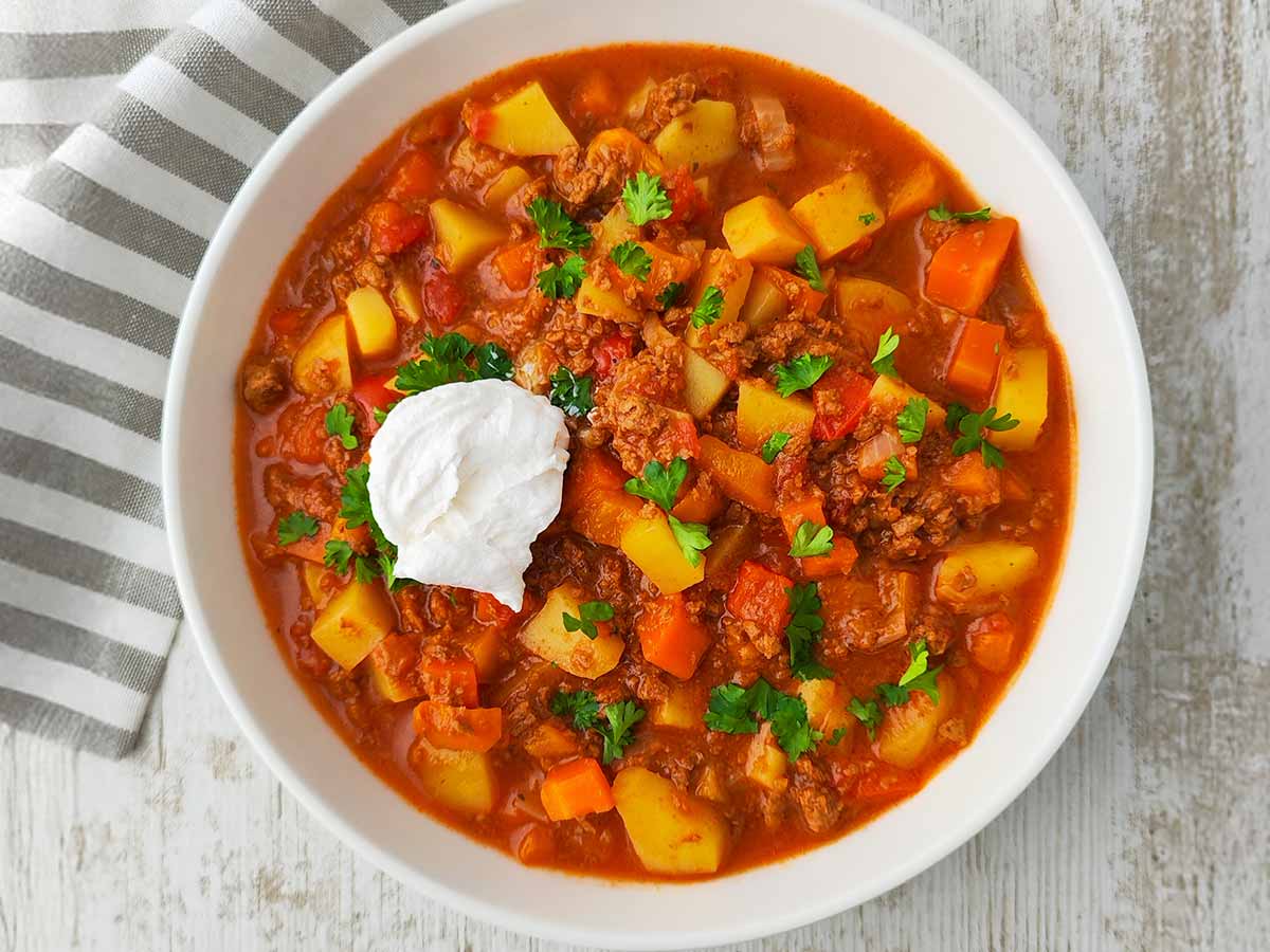 Teller mit veganem Bauerntopf mit veganem Hackfleisch, Kartoffeln, Möhren und Paprika auf hellem Holztisch.