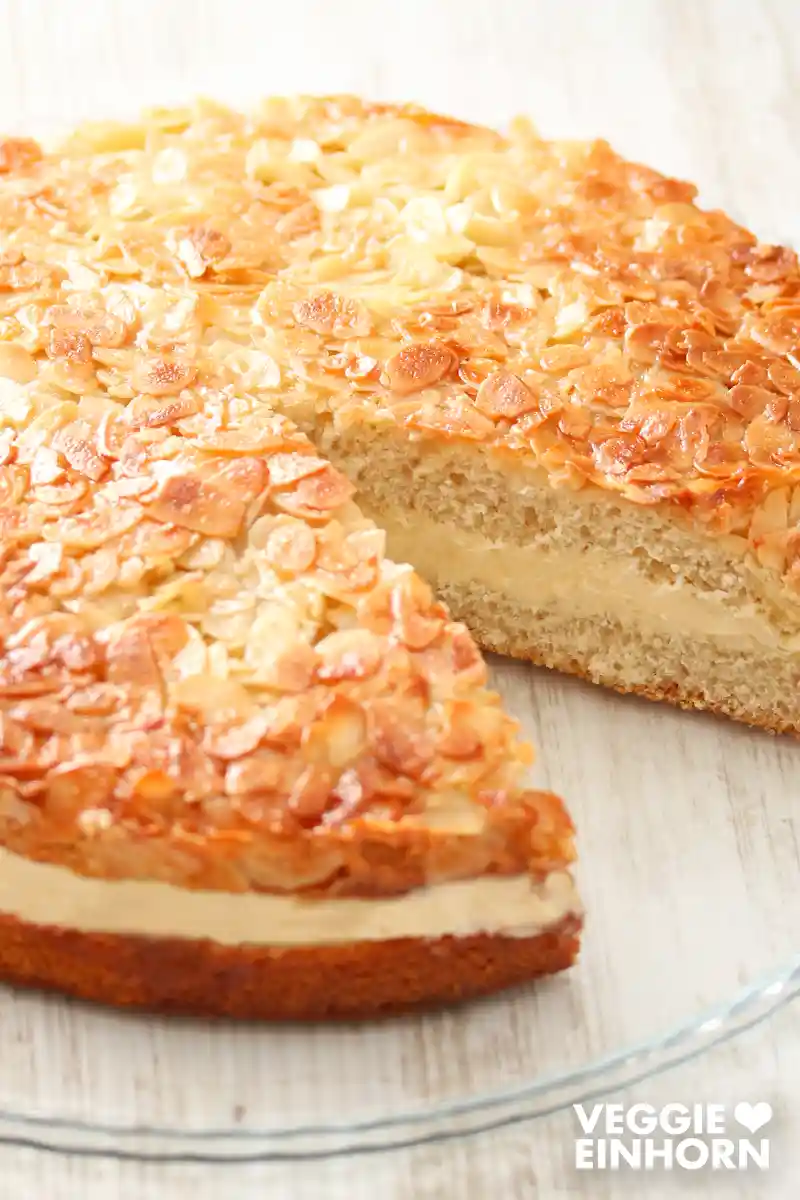Ein angeschnittener veganer Bienenstich auf einer runden Kuchenplatte aus Glas.