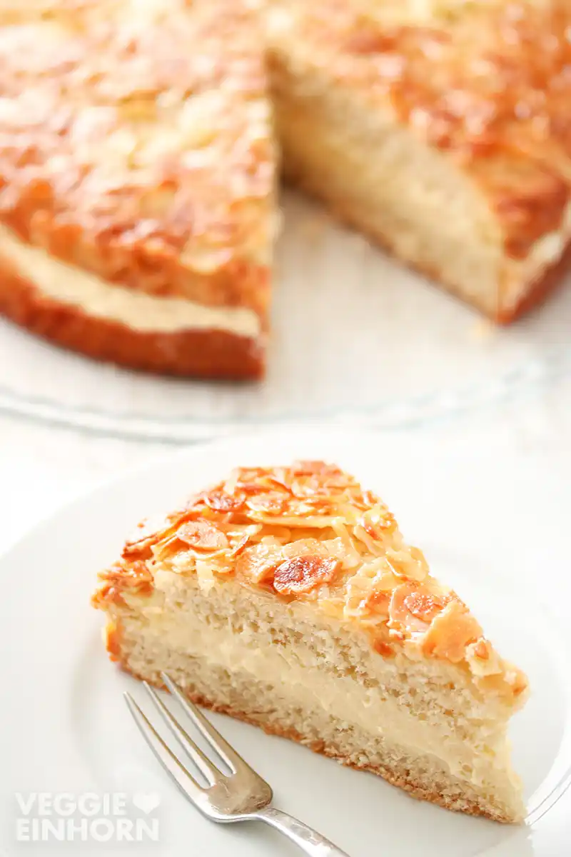 Ein Stück veganer Bienenstich auf einem Kuchenteller, im Hintergrund der angeschnittene Kuchen.