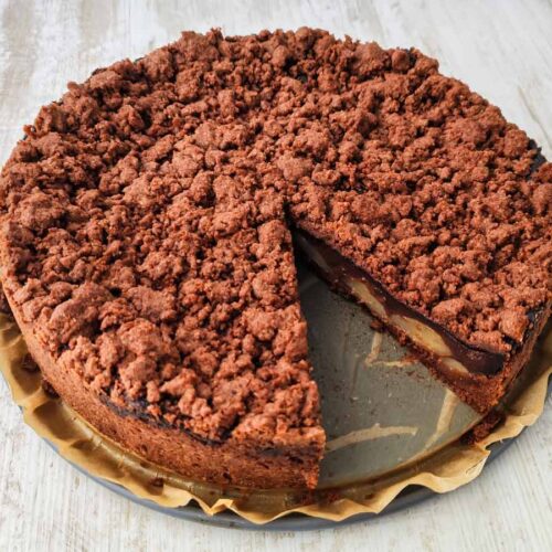 Ein runder veganer Schoko-Birnenkuchen mit Streuseln und Schokopudding, bei dem bereits ein Stück herausgeschnitten wurde.
