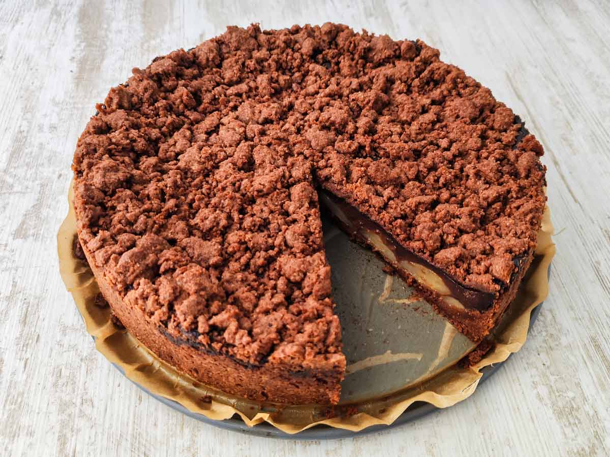 Ein runder veganer Schoko-Birnenkuchen mit Streuseln und Schokopudding, bei dem bereits ein Stück herausgeschnitten wurde.