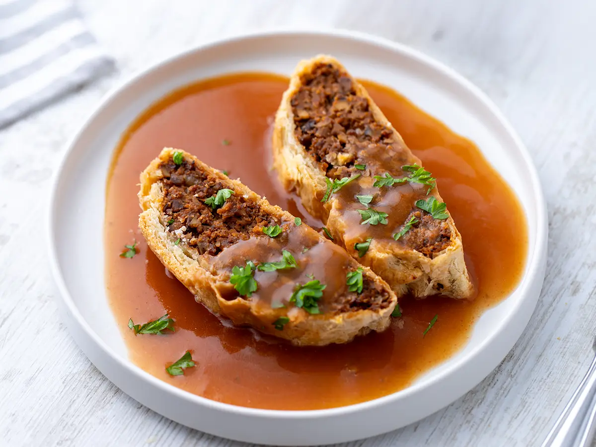 Zwei Scheiben veganer Braten im Blätterteig mit veganer brauner Sauce und Petersilie auf einem weißen Teller.