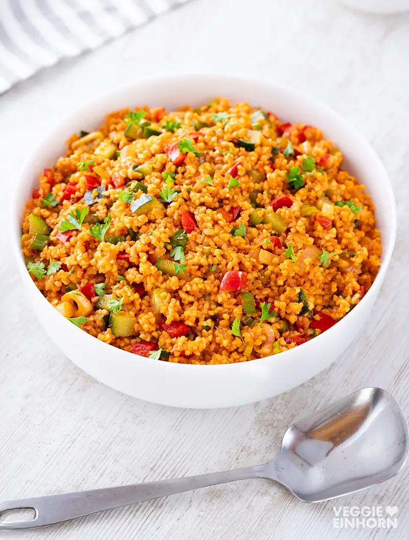 Veganer Bulgursalat in einer weißen Schüssel auf hellem Untergrund mit Löffel daneben.
