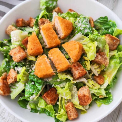 Ein weißer Teller mit veganem Caesar Salat aus Römersalat, Croutons, veganem Parmesan und veganen Nuggets.