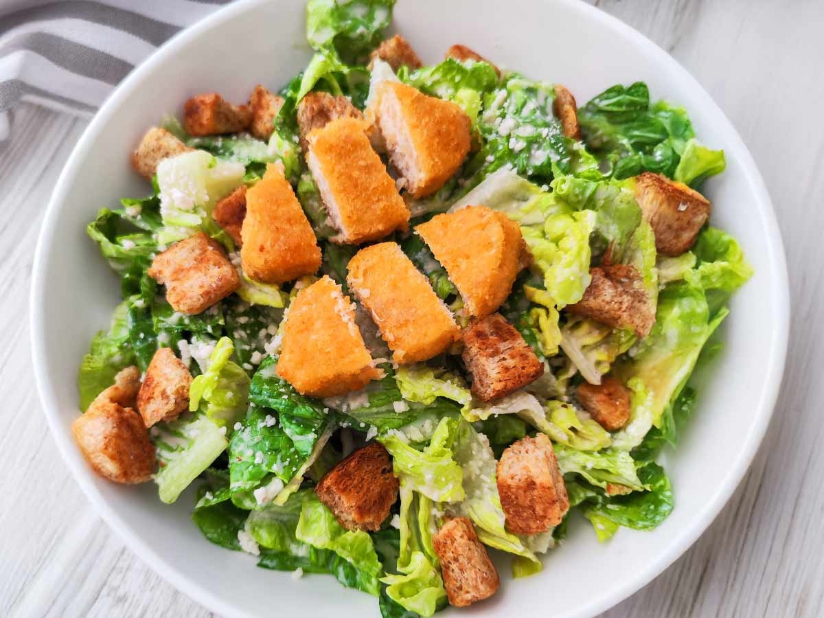 Ein weißer Teller mit veganem Caesar Salat aus Römersalat, Croutons, veganem Parmesan und veganen Nuggets.