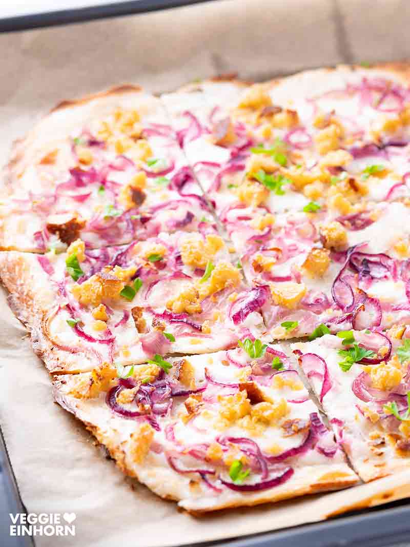 Veganer Flammkuchen auf einem Backblech mit Backpapier, belegt mit veganer Crème fraîche, roten Zwiebeln, Räuchertofu und Petersilie.