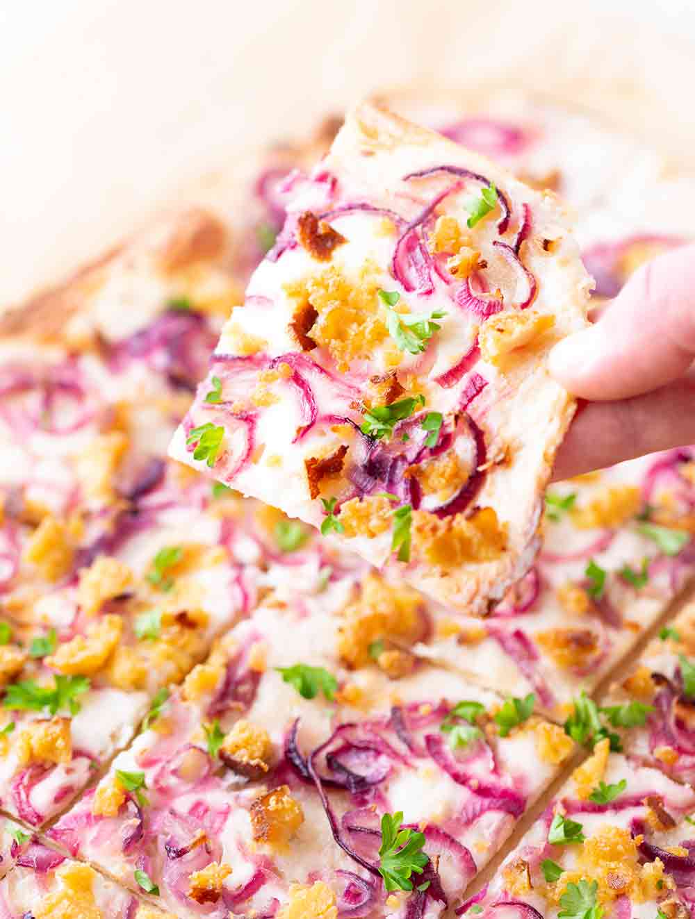 Ein Stück veganer Flammkuchen wird von einer Hand gehalten. Sichtbar sind der knusprige Teigboden und der Belag aus veganer Crème fraîche, roten Zwiebeln, Räuchertofu und frischer Petersilie.