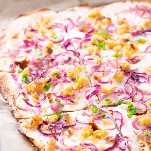 Veganer Flammkuchen mit dünnem knusprigen Boden, bestrichen mit veganer Crème fraîche, belegt mit roten Zwiebeln, Räuchertofu-Stückchen und frischer Petersilie.