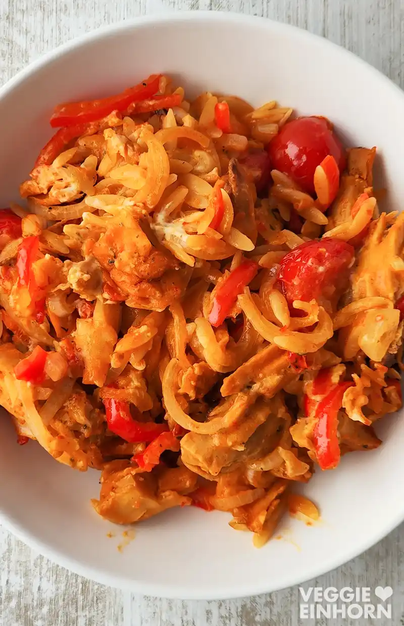 Veganer Gyrosauflauf mit Kritharaki, veganem Feta, roten Paprikastreifen, Zwiebeln und Kirschtomaten auf einem weißen Teller.