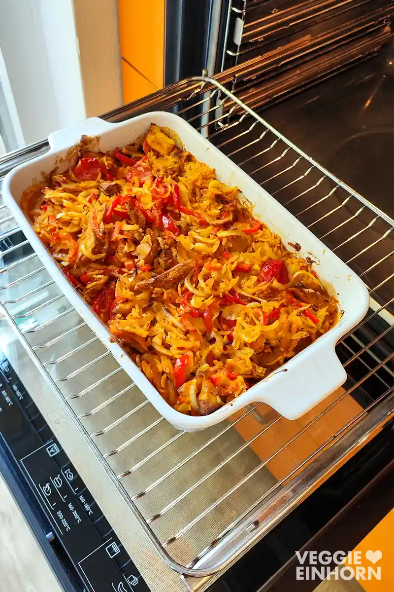 Kritharaki-Auflauf mit veganem Gyros, veganem Feta, Paprika, Zwiebeln und Tomaten in einer Auflaufform auf dem Backofenrost.