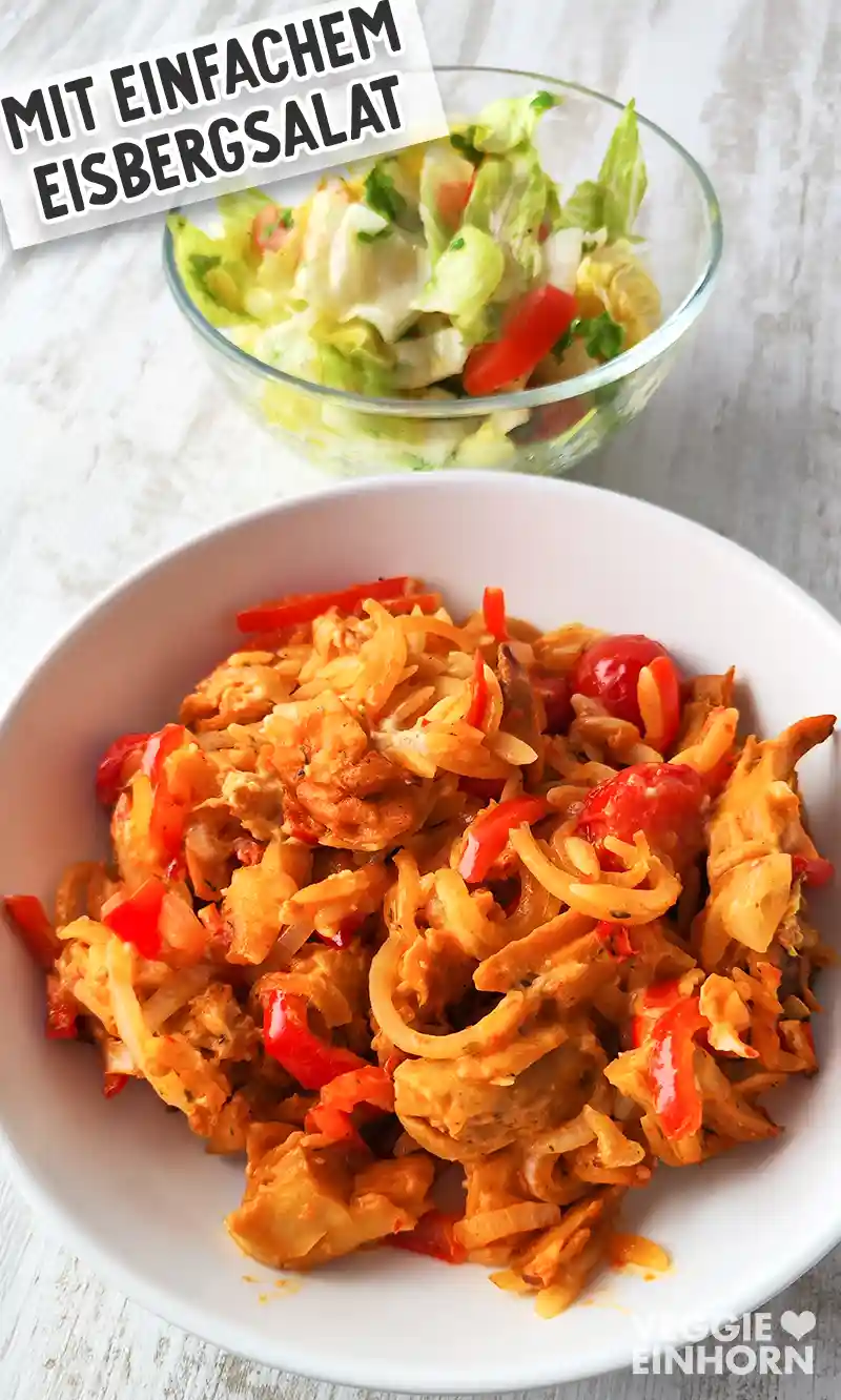 Veganer Gyrosauflauf mit Paprika, Zwiebeln, Kirschtomaten und Kritharaki auf einem weißen Teller, im Hintergrund ein Glasschälchen mit einfachem Eisbergsalat mit Tomaten und Petersilie.