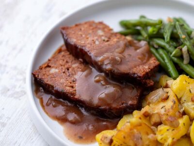 Vegane Scheiben Hackbraten aus Kidneybohnen und Linsen mit Soße, grünen Bohnen und Bratkartoffeln, serviert auf einem weißen Teller.