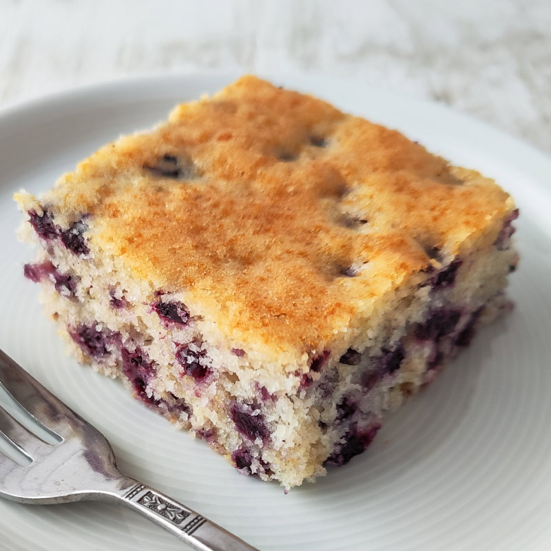 Ein Stück veganer Heidelbeer-Blechkuchen auf einem Teller, daneben eine Kuchengabel.