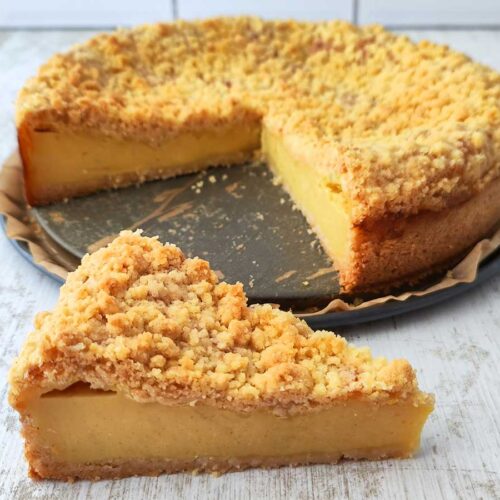 Ein veganer Käsekuchen mit goldbraunen Streuseln auf einer Springform, vorne liegt ein angeschnittenes Stück auf dem Tisch.