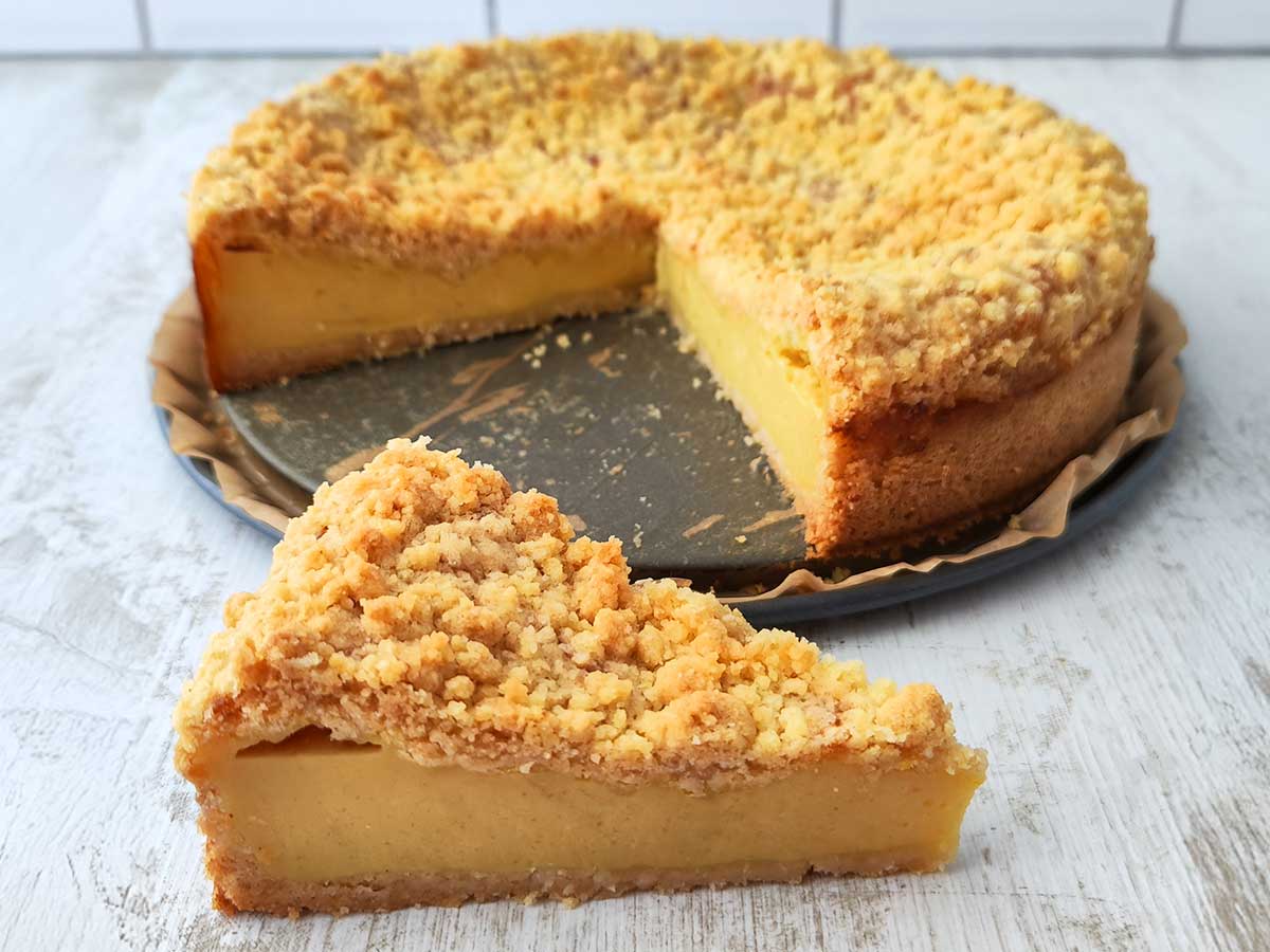 Ein veganer Käsekuchen mit goldbraunen Streuseln auf einer Springform, vorne liegt ein angeschnittenes Stück auf dem Tisch.