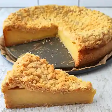 Ein veganer Käsekuchen mit goldbraunen Streuseln auf einer Springform, vorne liegt ein angeschnittenes Stück auf dem Tisch.