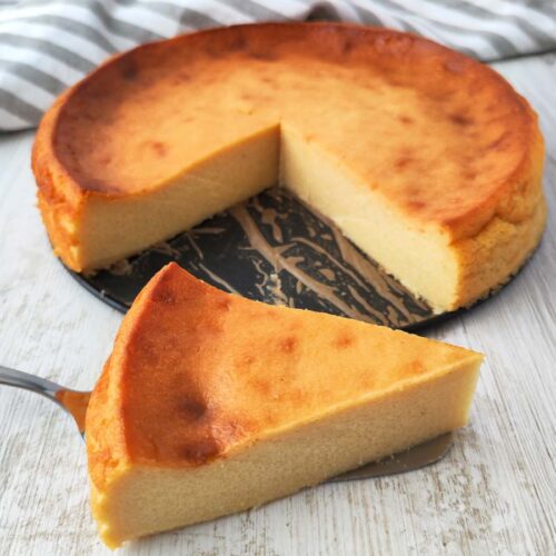 Ein veganer Käsekuchen ohne Boden mit leicht gebräunter Oberfläche, ein Stück liegt davor auf einem Tortenheber.