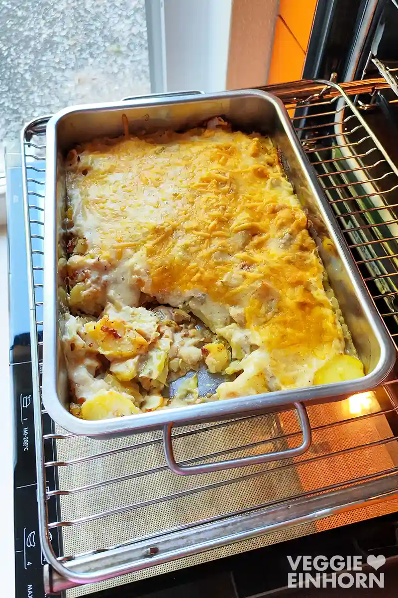 Auflaufform mit frisch gebackenem veganen Kartoffel-Spitzkohl-Auflauf auf dem Backofenrost vor dem geöffneten Backofen.
