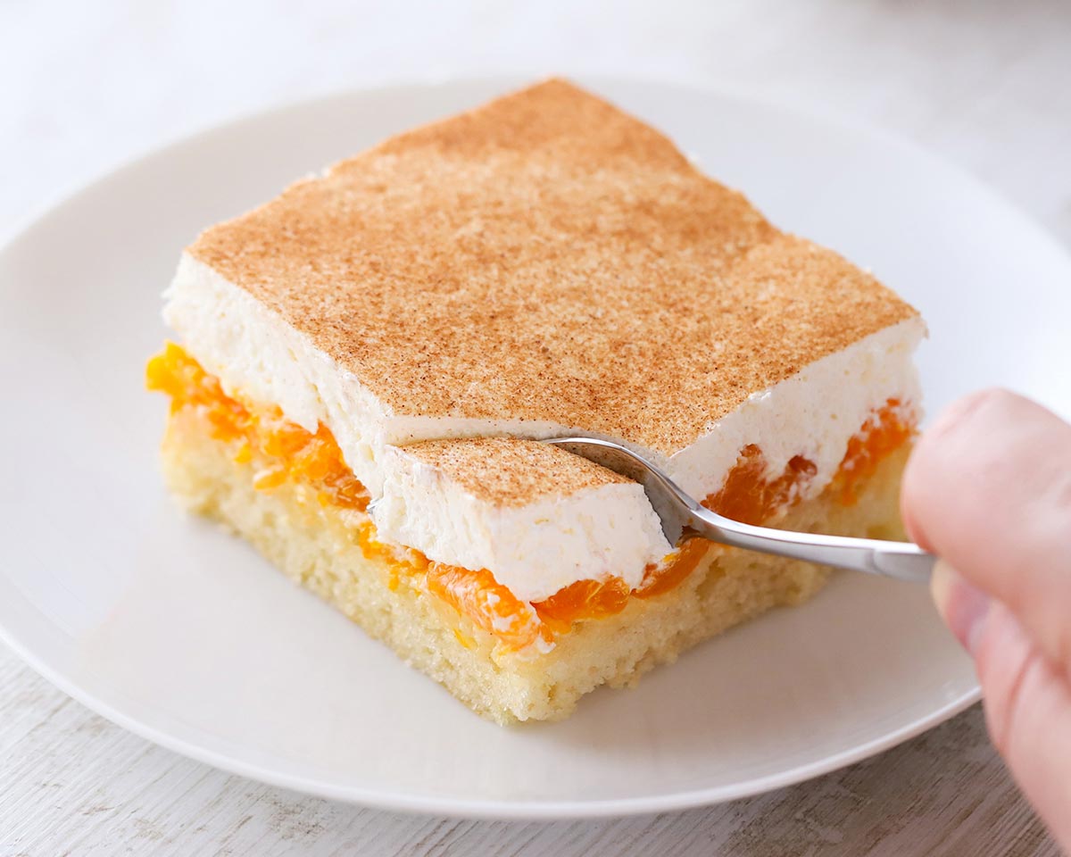 Ein Stück veganer Mandarinen-Schmand-Kuchen auf einem weißen Teller wird mit einer Gabel geteilt.