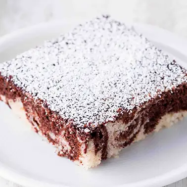 Ein Stück veganer Marmorblechkuchen, bestreut mit Puderzucker, serviert auf einem weißen Teller.