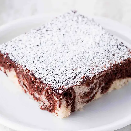 Ein Stück veganer Marmorkuchen vom Blech mit heller und dunkler Teigschicht, bestäubt mit Puderzucker, serviert auf einem weißen Teller.
