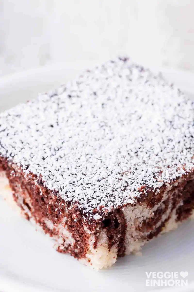 Nahaufnahme eines Stücks veganen Marmorblechkuchens mit heller und dunkler Marmorierung, großzügig mit Puderzucker bestreut, serviert auf einem weißen Teller.