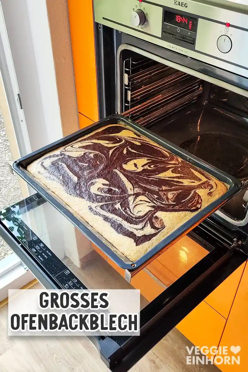 Ein großes Blech veganer Marmorblechkuchen mit heller und dunkler Marmorierung wird aus einem geöffneten Backofen gezogen.