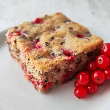 Ein Stück veganer Johannisbeerkuchen mit Nüssen und Schokolade liegt auf einem weißen Teller. Daneben liegen frische rote Johannisbeeren.