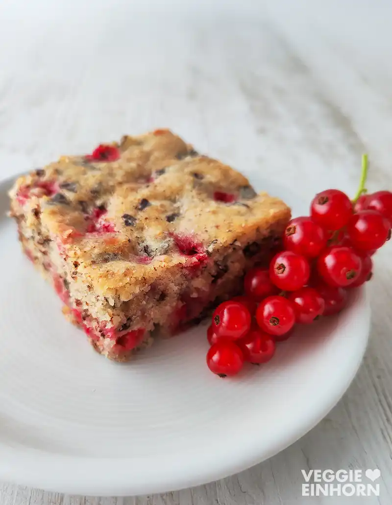 Ein Stück veganer Johannisbeerkuchen mit Nüssen und Schokoraspeln liegt auf einem weißen Teller. Daneben befindet sich eine kleine Rispe frischer roter Johannisbeeren. Der Teller steht auf einem hellen Holztisch.