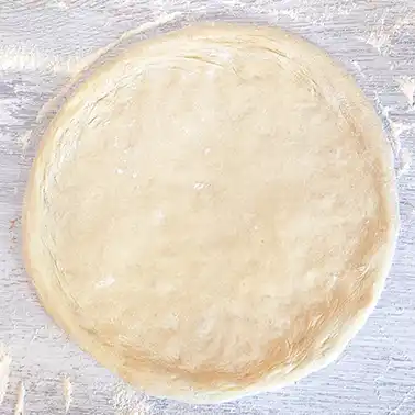 Ein runder, ungebackener Pizzateig liegt auf einer bemehlten Holzoberfläche.