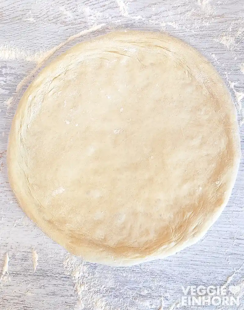 Ein runder, ungebackener Pizzateig mit einem Rand liegt auf einer bemehlten Arbeitsfläche.