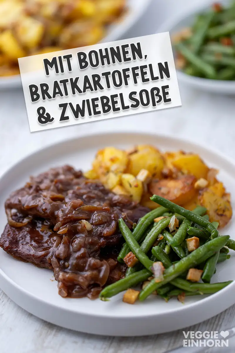 Veganer Zwiebelrostbraten mit Bohnen, Bratkartoffeln und Zwiebelsauce auf einem weißen Teller.