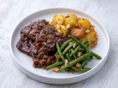 Nahaufnahme von veganem Rostbraten aus Soja Big Steaks mit Zwiebelsauce, Bratkartoffeln und grünen Bohnen.