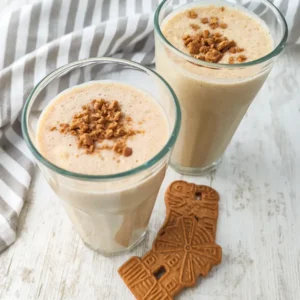 Zwei Gläser veganer Spekulatius-Milchshake stehen auf einem hellen Tisch. Daneben liegen Spekulatius Kekse und ein gestreiftes Küchentuch.