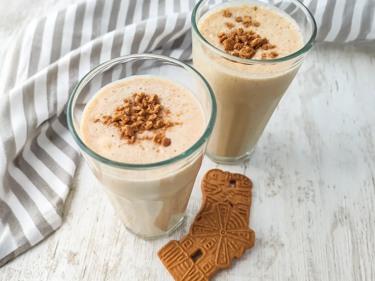 Zwei Gläser veganer Spekulatius-Milchshake stehen auf einem hellen Tisch. Daneben liegen Spekulatius Kekse und ein gestreiftes Küchentuch.